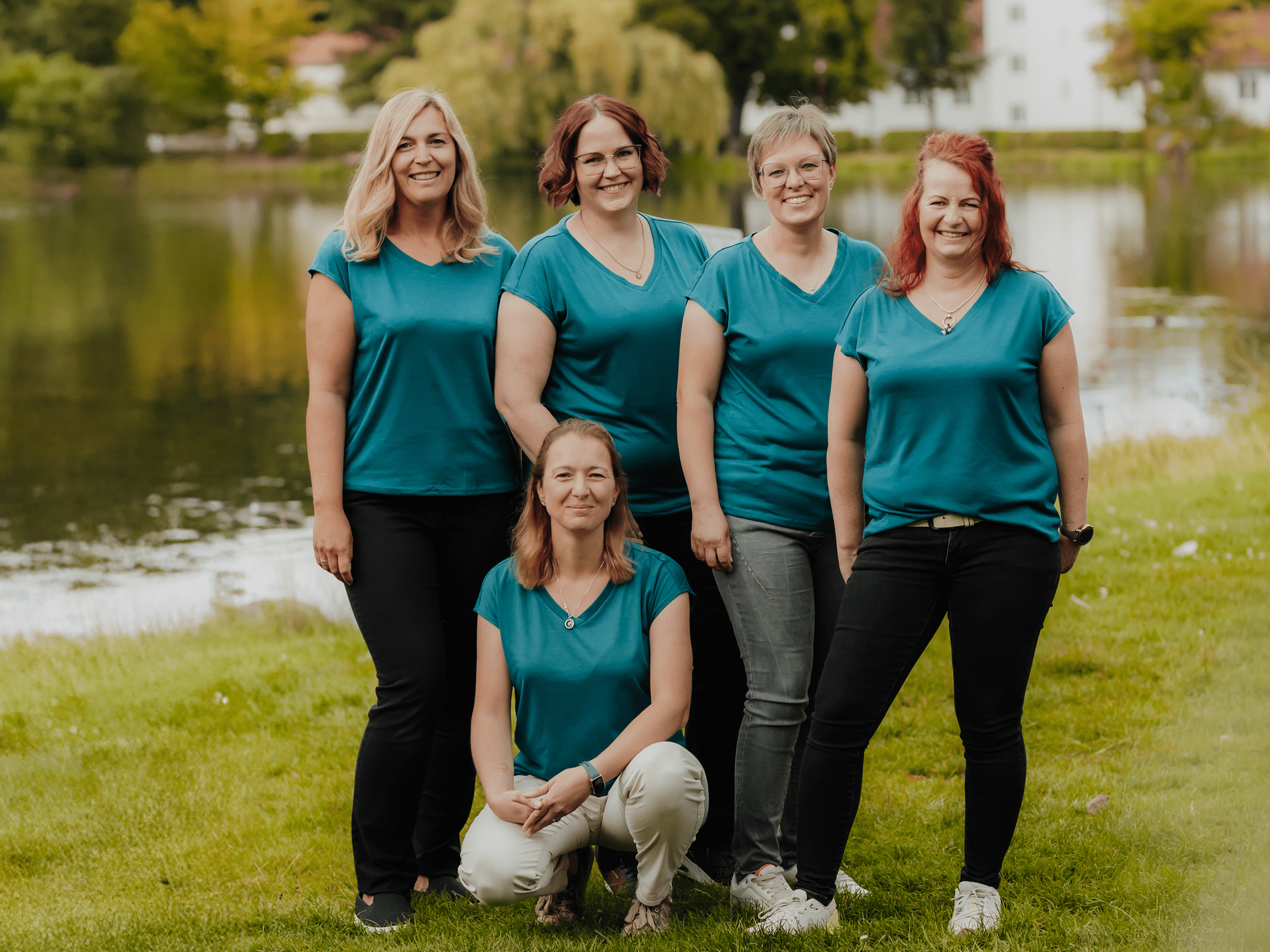 Fünf Frauen stehen und knien auf einer Wiese am Ufer eines Sees. Sie tragen alle türkisfarbene T-Shirts und dunkle Hosen und lächeln in die Kamera. Im Hintergrund sind Wasser, Bäume und einige helle Gebäude zu sehen.